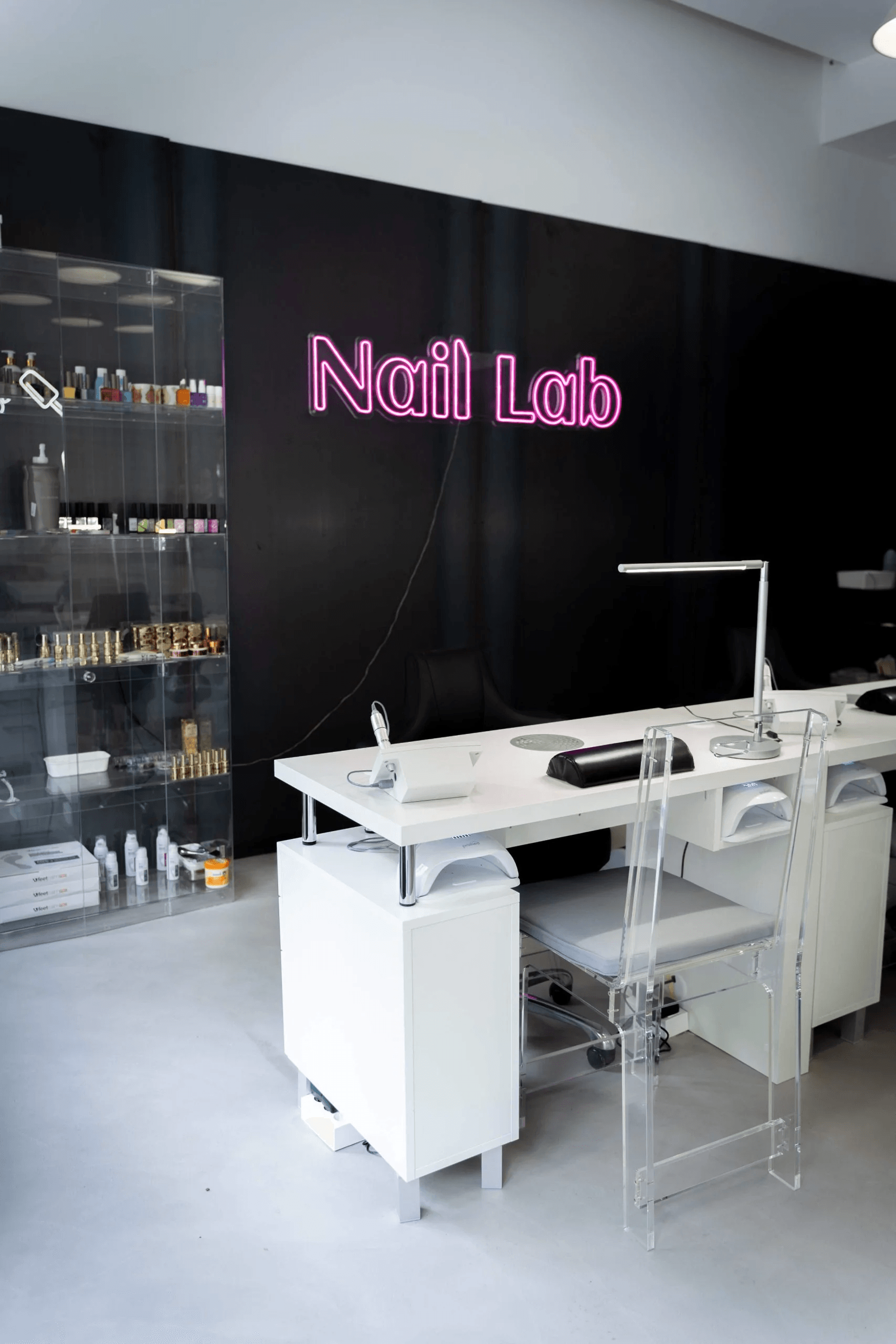 White nail work desk, transparent chair, a hot pink Nail Lab logo neon sign visible in the background on a black wall